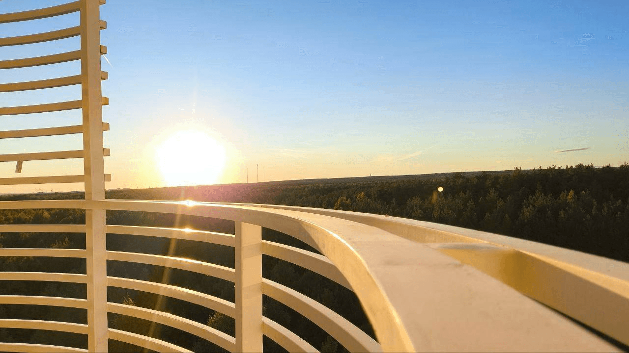 Futuristic white building with curved design, large windows, and glowing orange interior at sunset. Reflecting pool, palm trees, calm atmosphere.