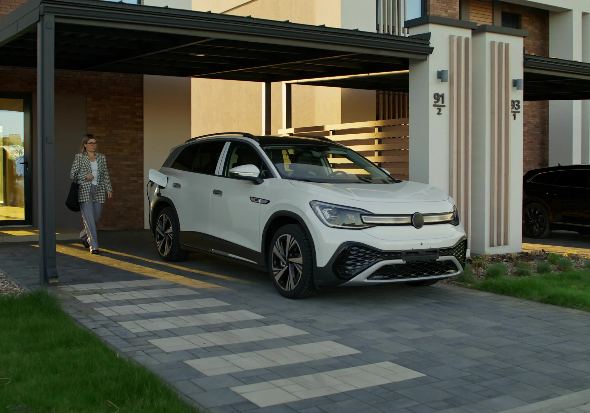 A modern white SUV is parked in a stylish driveway under a sleek carport. A woman in business attire walks towards the vehicle, conveying a professional tone.