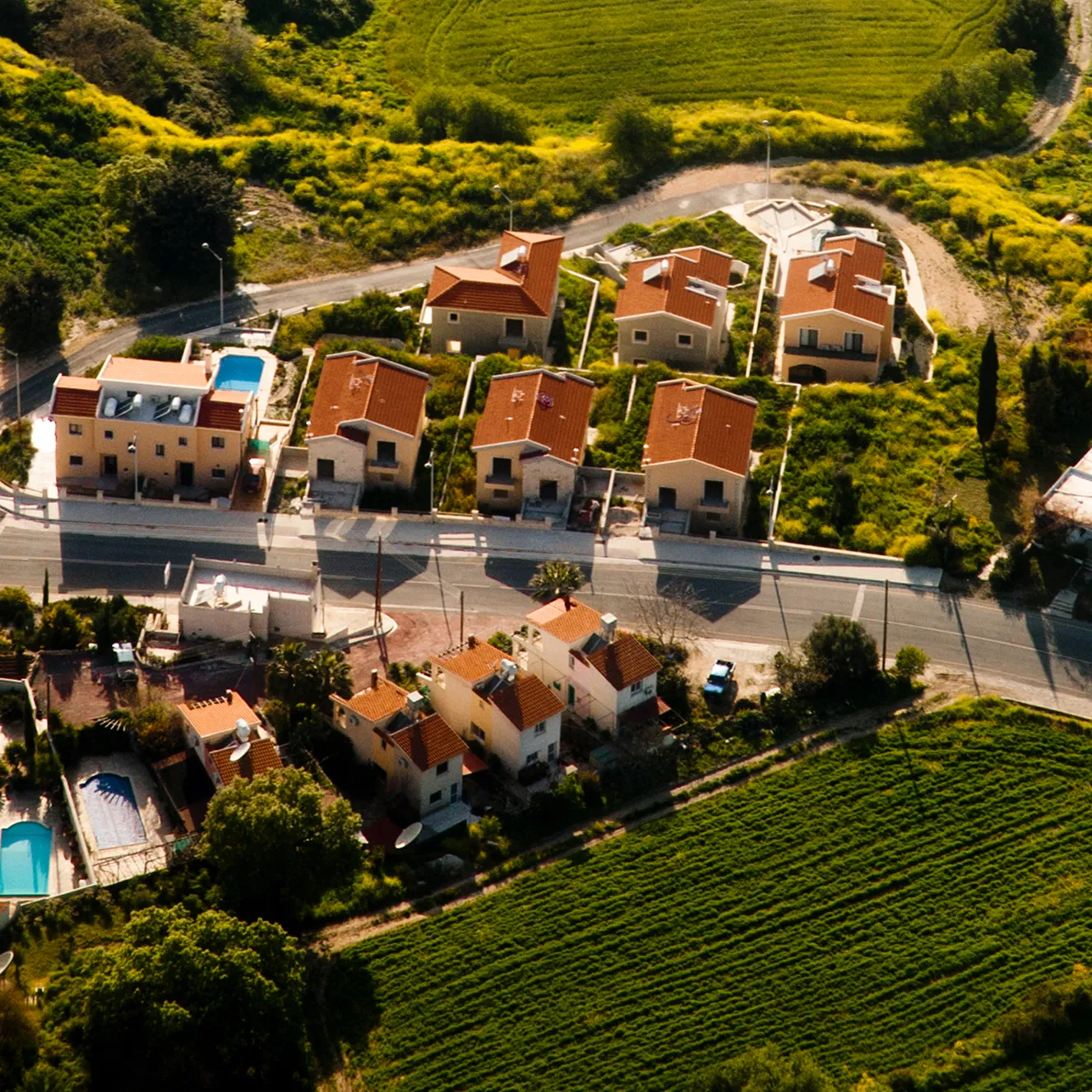 Aerial view of a small neighborhood with red-roofed houses nestled among lush green fields and winding roads. The scene feels peaceful and idyllic.